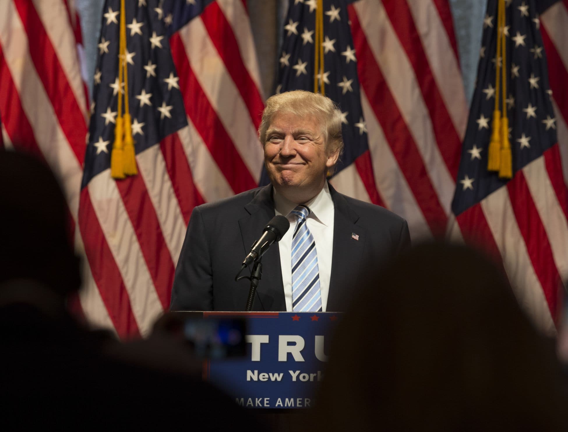 Donal Trump smiling on a podium with American flags behind him