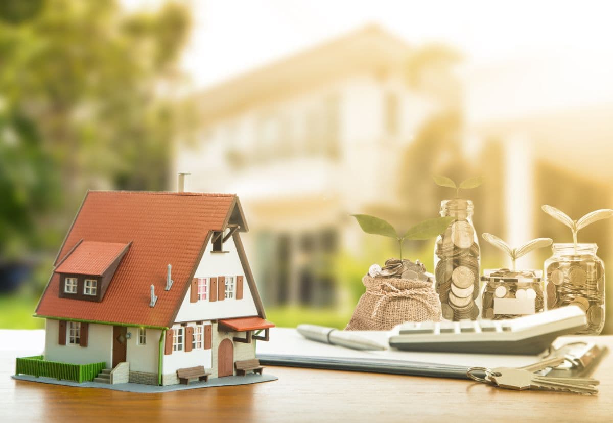 A model house on a table next to a stack of papers, calculator and jars of coins with a seedling growing from them